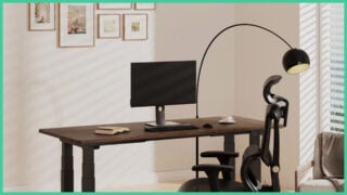 The FlexiSpot E7 Plus with a PC monitor, keyboard, mouse, a cup and saucer, and an open book, with framed pictures on the wall.