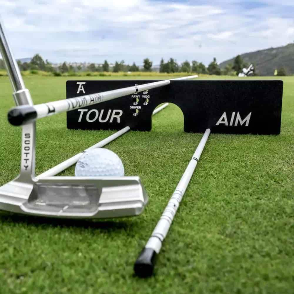 A golf putter, a golf ball, and two alignment rods are set up on grass with a black Tour Aim training aid in the background&mdash;an ideal setup for practicing golf alignment and finding the best deals under $50. Trees and hills can be seen in the distance.