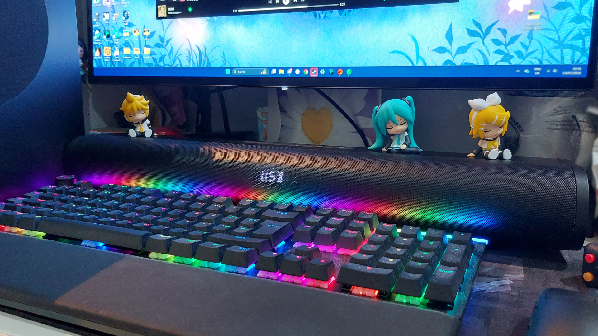 The Creative Stage Pro sits on a desk before a monitor and behind an RGB keyboard. Miniature Vocaloid figurines sit along the top of the soundbar. The soundbar's LED display shows the device is in USB mode.