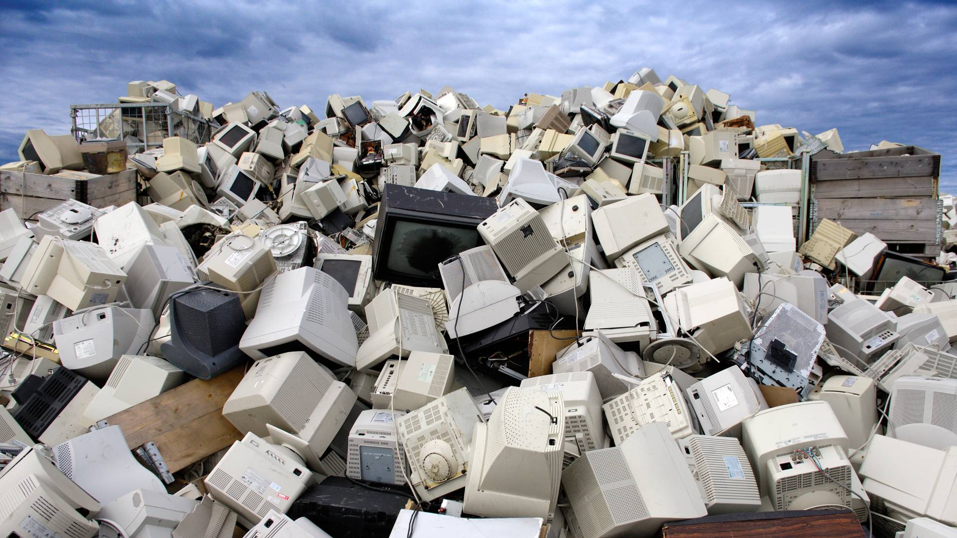 A recycling plant for tellies and monitors in Norway. A large pile of CRT monitors and screens in various states of disrepair are haphazardly piled on top of eachother. A partially cloudy blue sky can be seen beyond this mountain of e-waste.