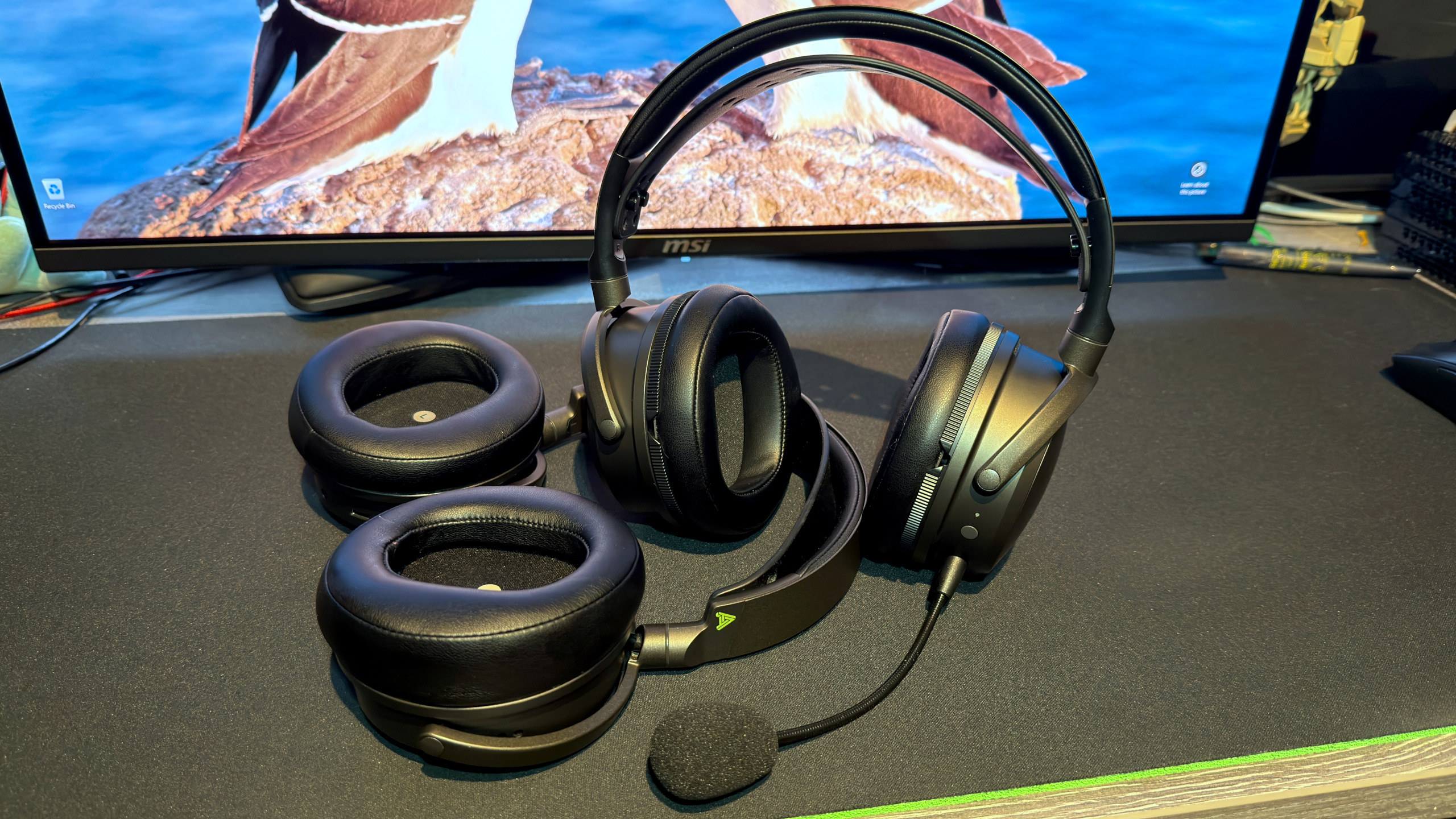 A photo of the Audeze Maxwell 2 wireless gaming headset, with a gaming monitor in the background, and the original Maxwell headset next to it