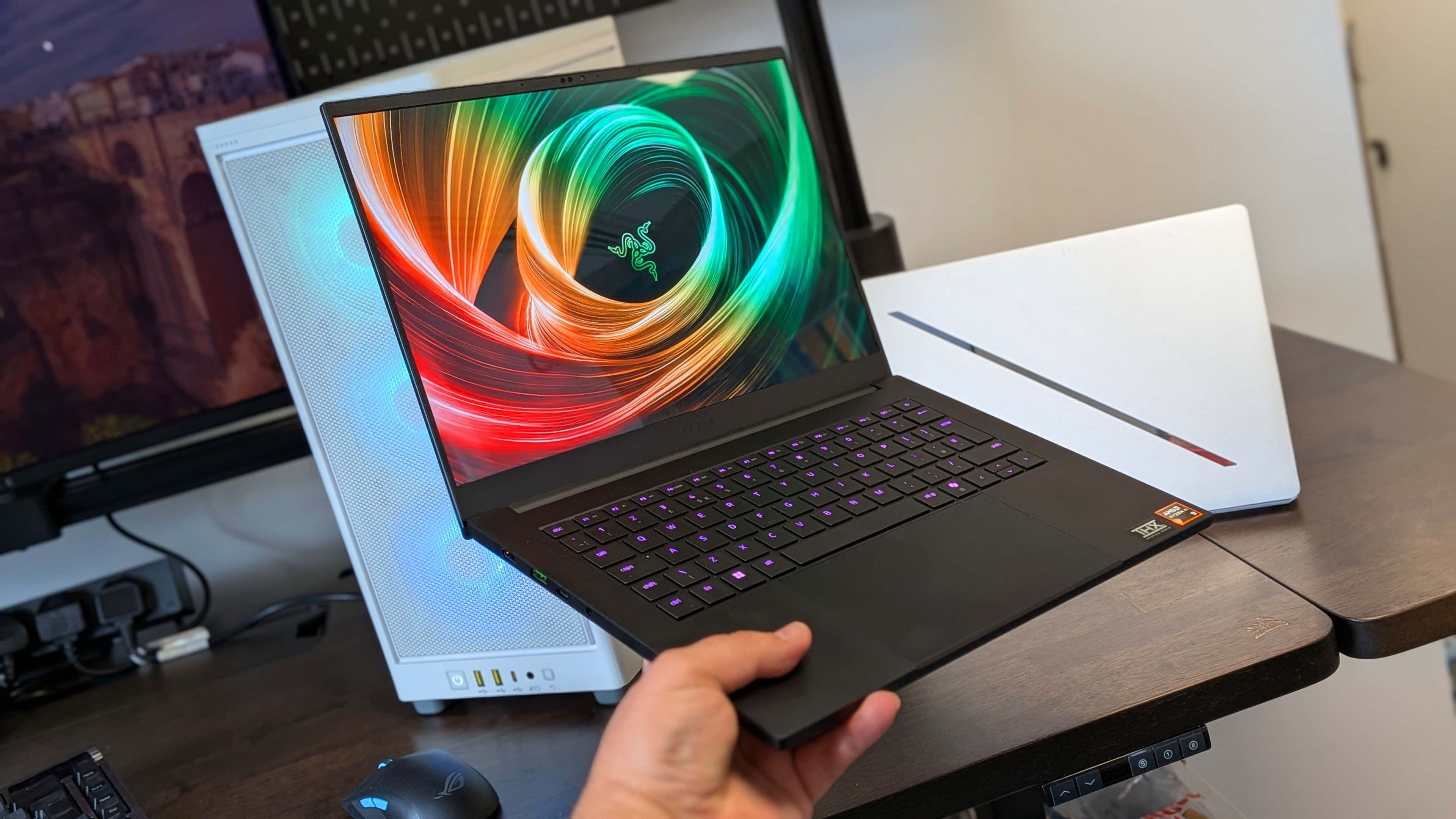 Razer Blade 14 gaming laptop held in one hand with a PC and an Asus ROG Zephyrus G14 in the background.